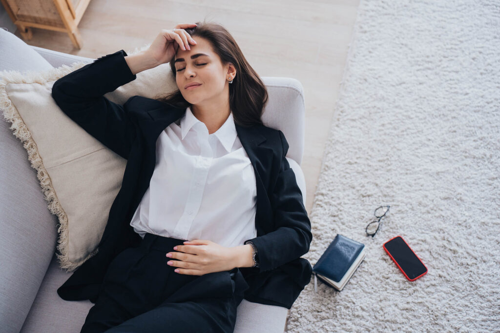 Eine Frau liegt völlig ermüdet auf dem Sofa, Telefon und Arbeitsgeräte noch neben ihr.